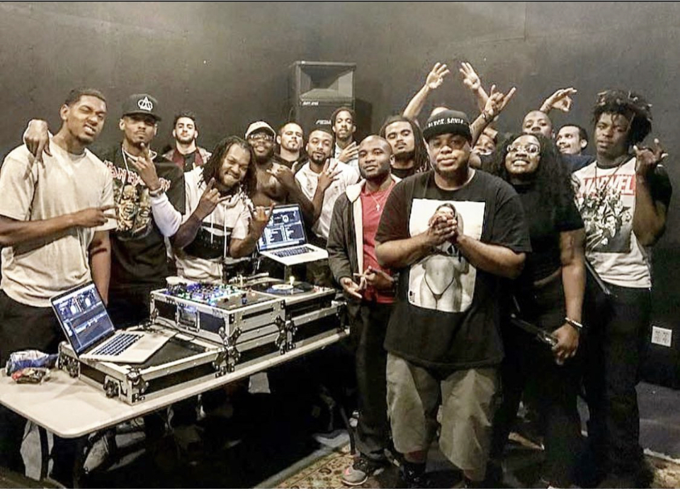 Group of people gathered around DJ equipment, some raising hands, in a dark indoor setting.