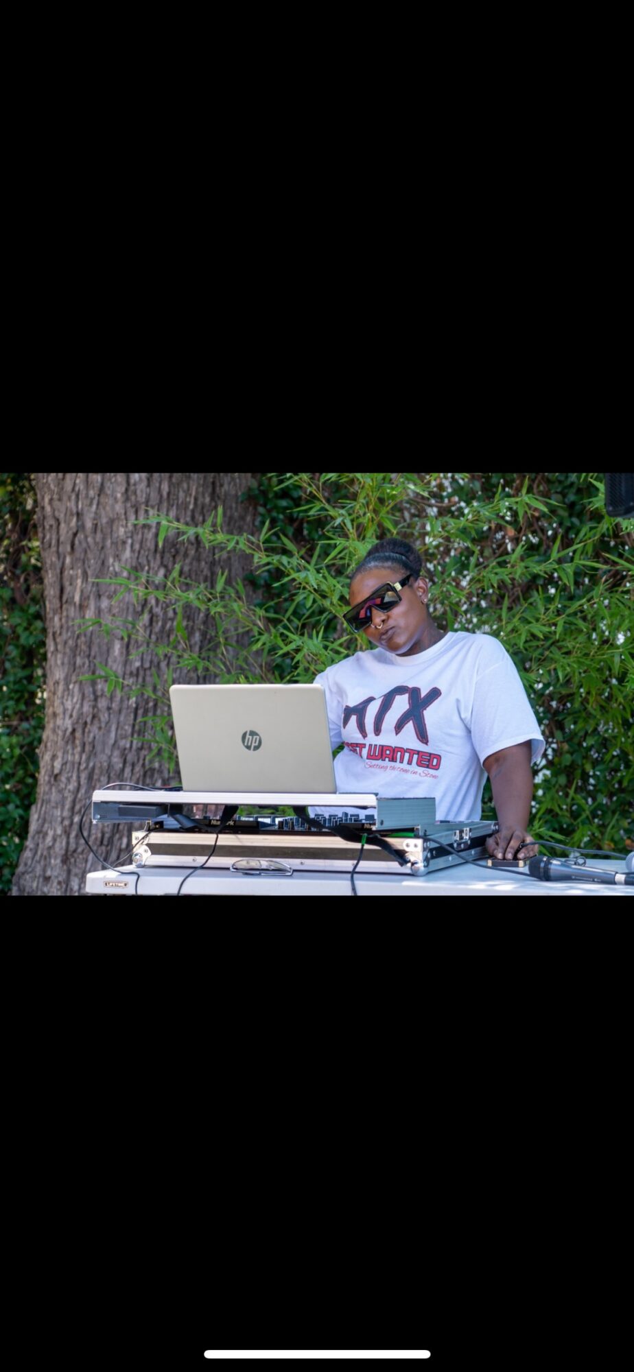 Person wearing sunglasses and a white T-shirt with a logo, standing outdoors with a laptop and DJ equipment, trees in background.