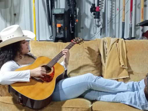 Person lying on a couch playing an acoustic guitar, wearing a white hat and light-colored clothing.