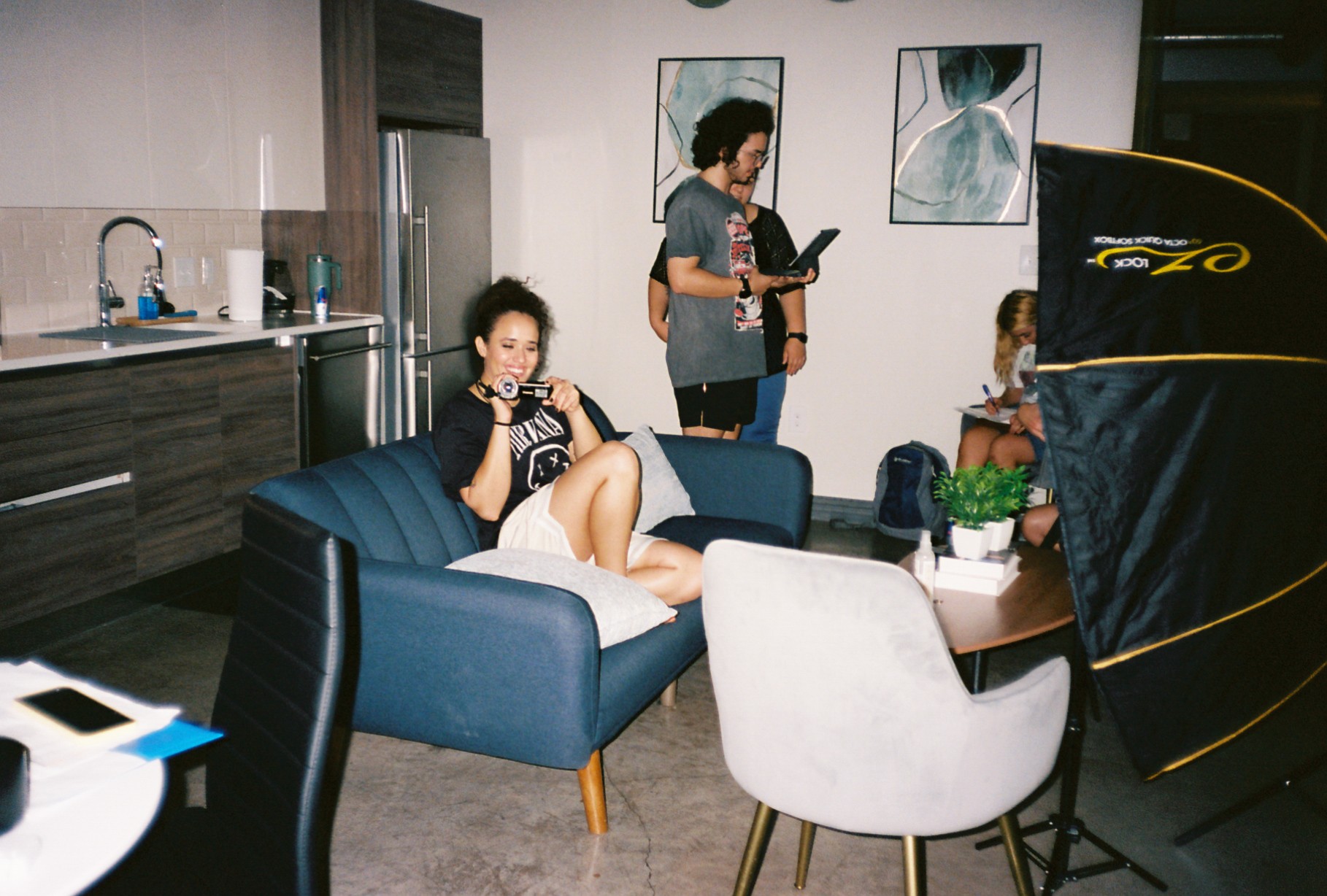 Person sitting on a blue sofa drinking from a cup, with two standing people and a white chair nearby in a modern room.