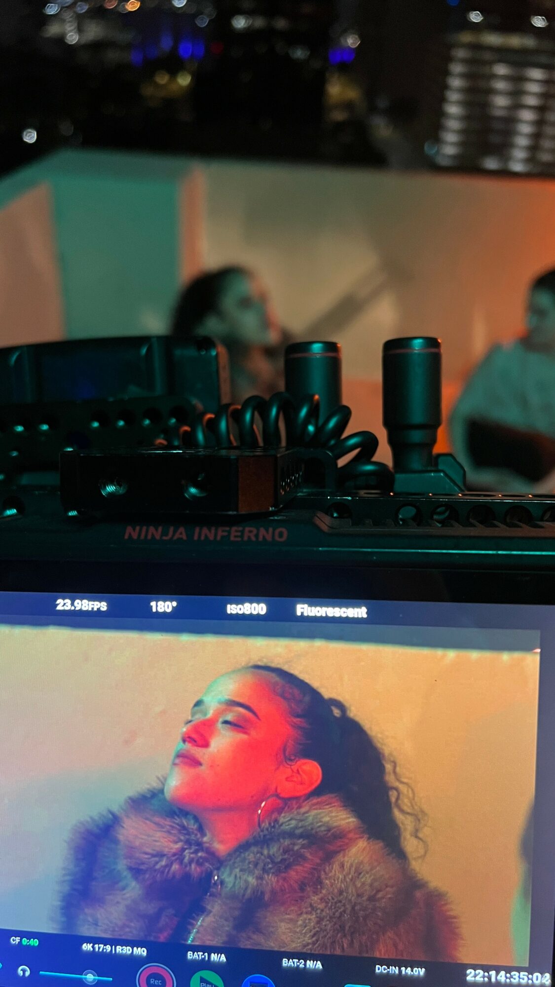 Close-up of a computer screen displaying a woman with curly hair and makeup, with a blurred background of people at a party.