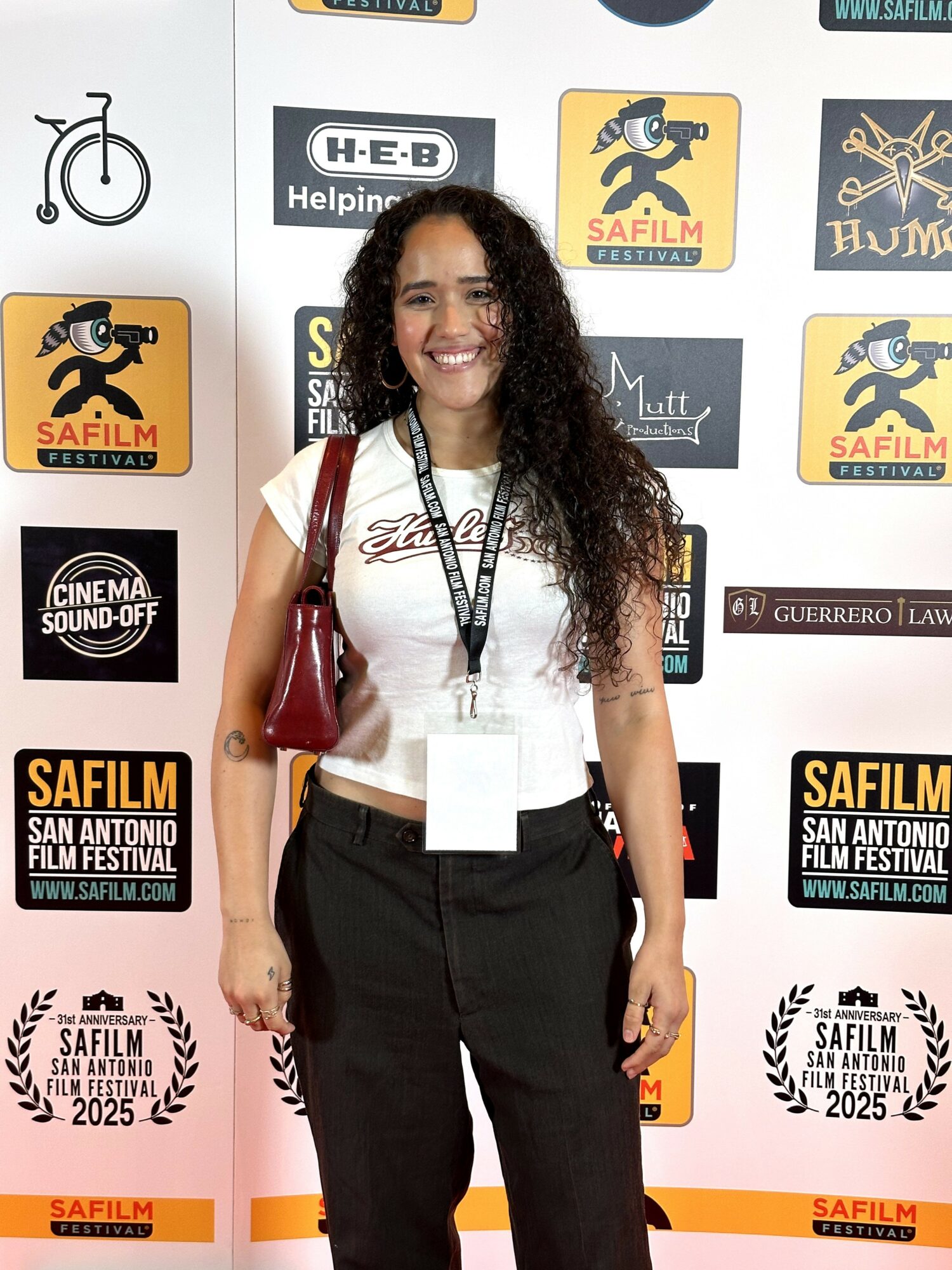 Woman with curly hair smiling, standing in front of a backdrop with festival logos, wearing a white top and black pants.