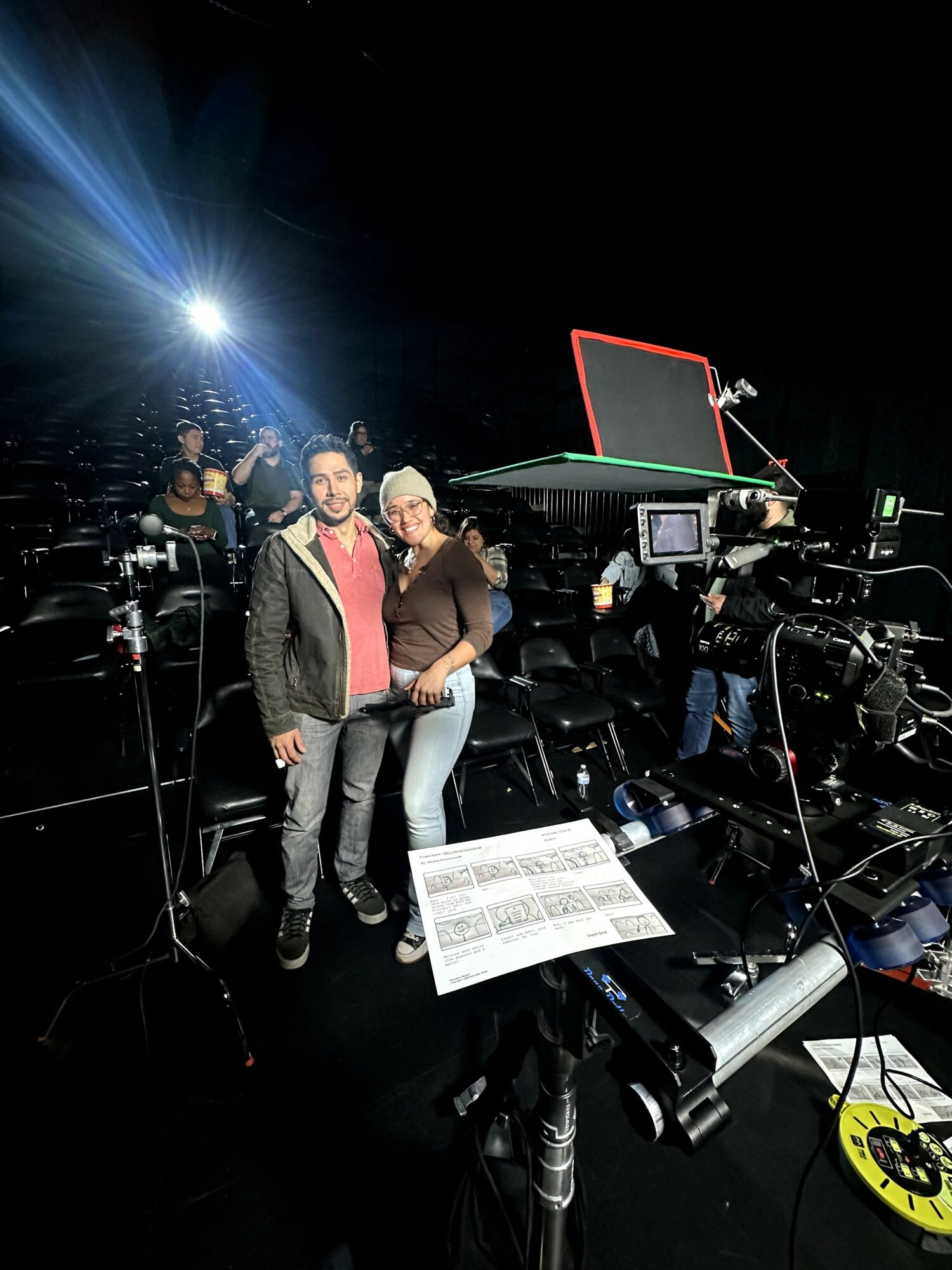 Two people stand together in a studio with cameras and equipment, bright lights, and a basketball hoop in the background.