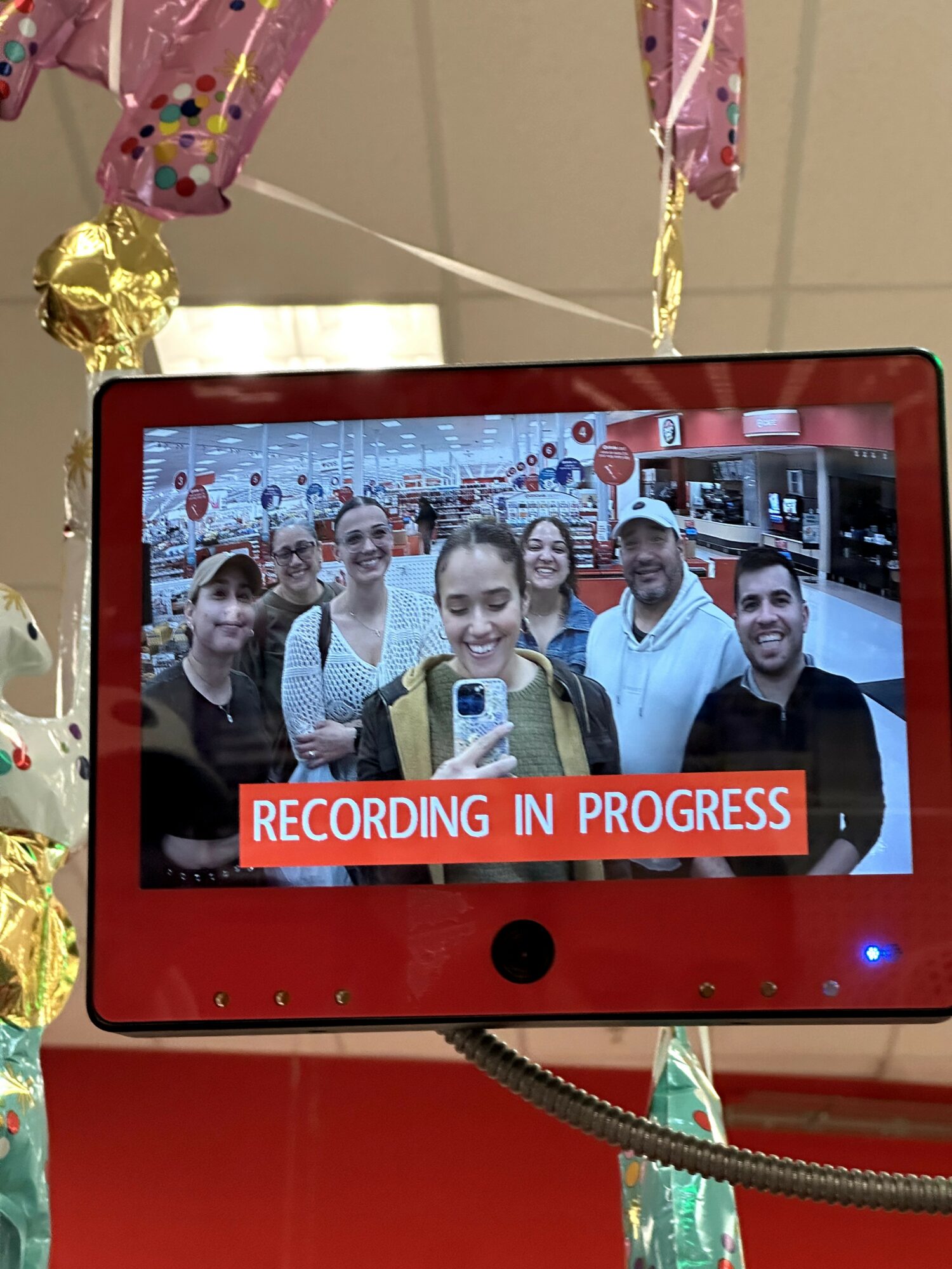 Group of six people taking a selfie in front of a screen with a red banner that says 'RECORDING IN PROGRESS'.