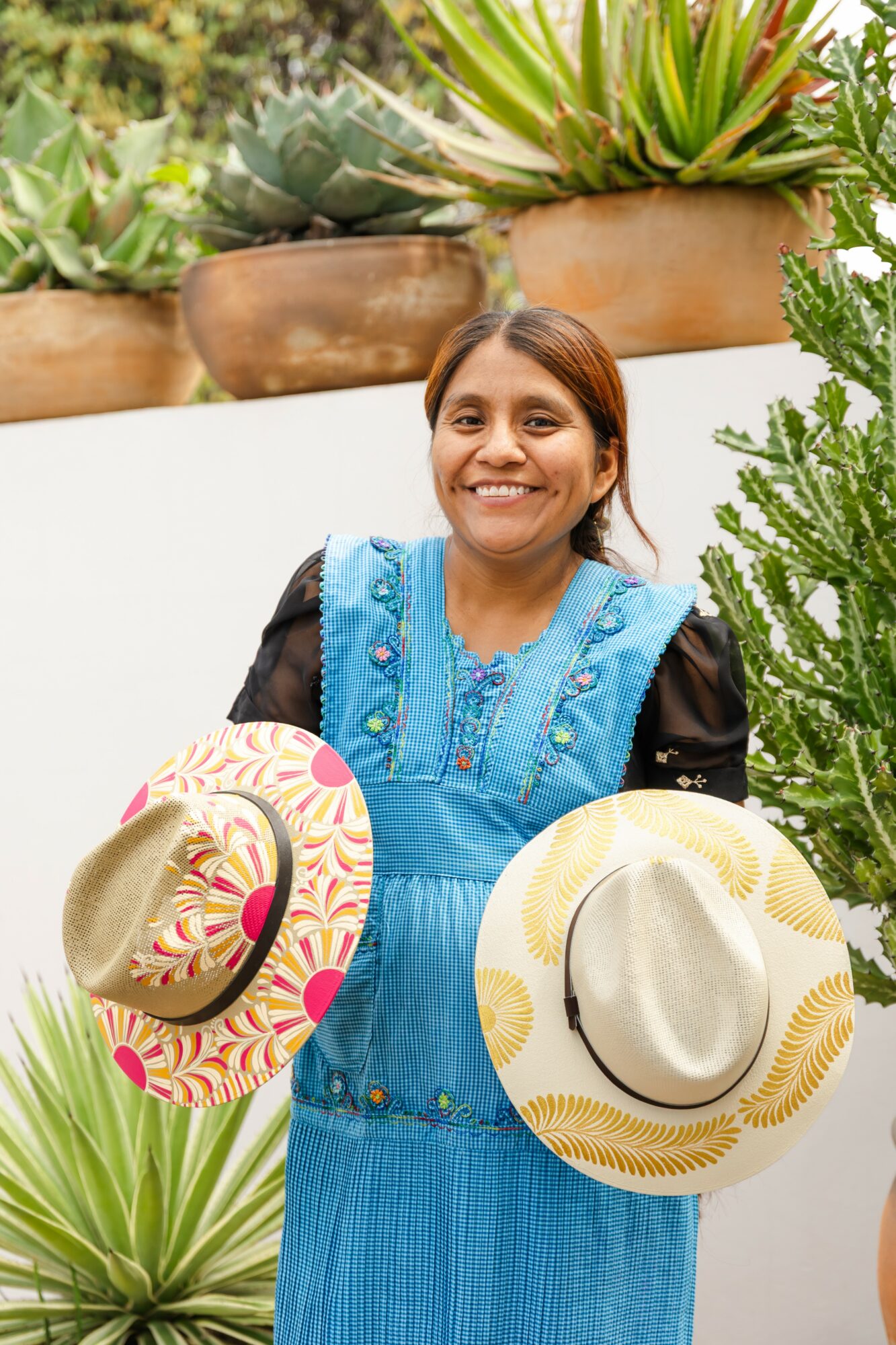 Smiling woman holding two hats outdoors with potted plants in background.