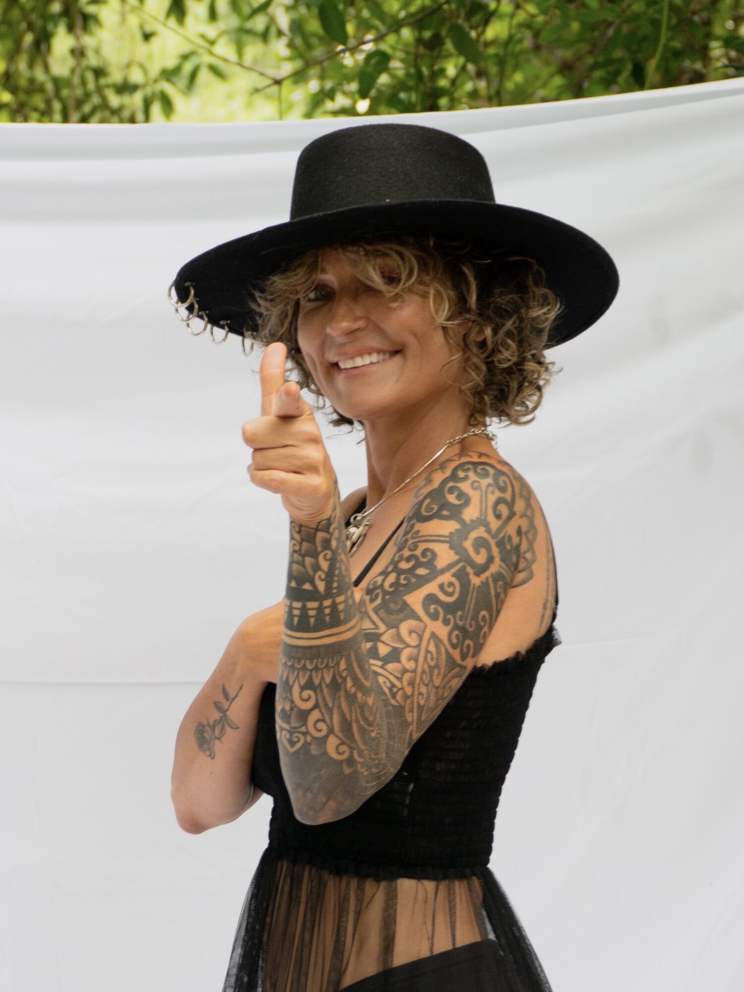 Woman with curly hair wearing a wide-brimmed hat, smiling, pointing, and showing tattoos on her arm and shoulder.