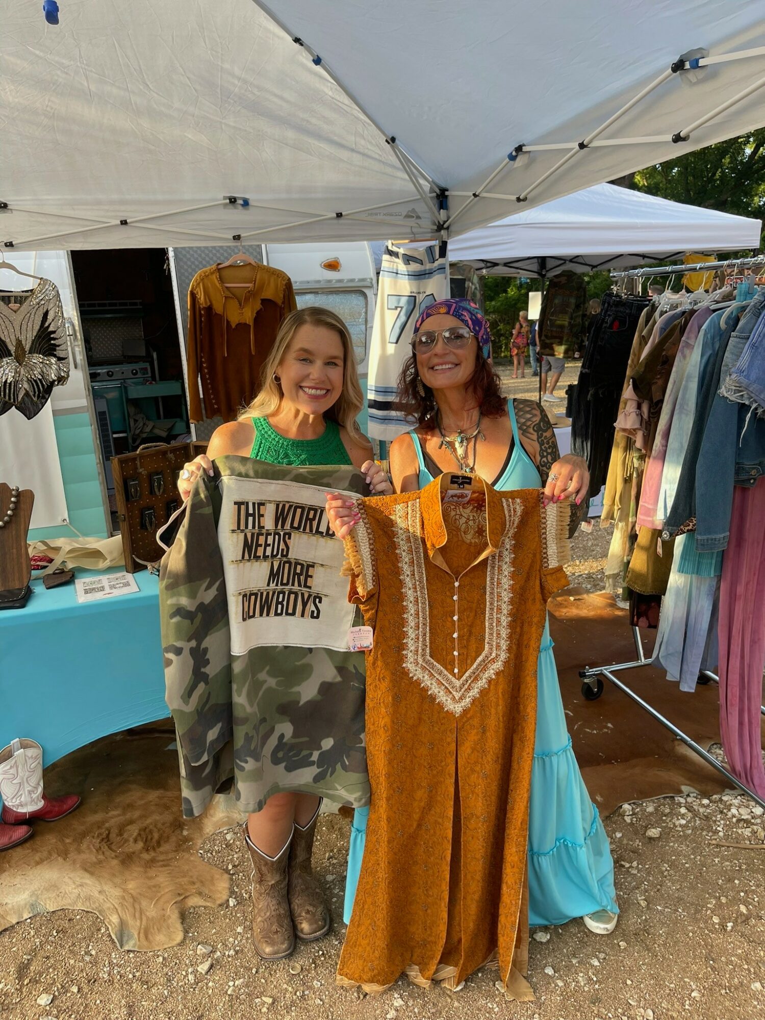 Two women smiling at a market stall with clothing, one holding a sign and the other holding a dress, under a white canopy.