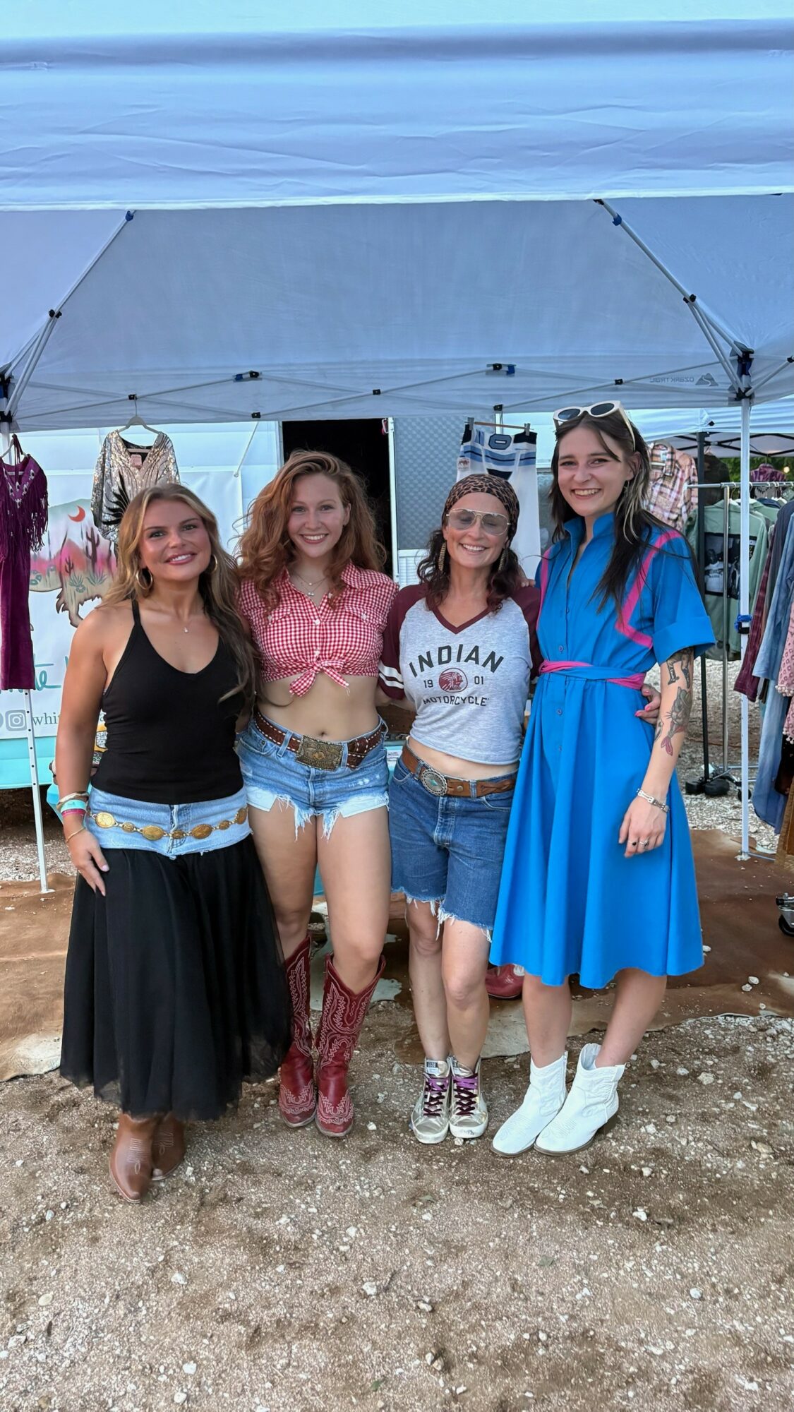 Four women stand outdoors under a tent, smiling, with a background of tents and outdoor setting.