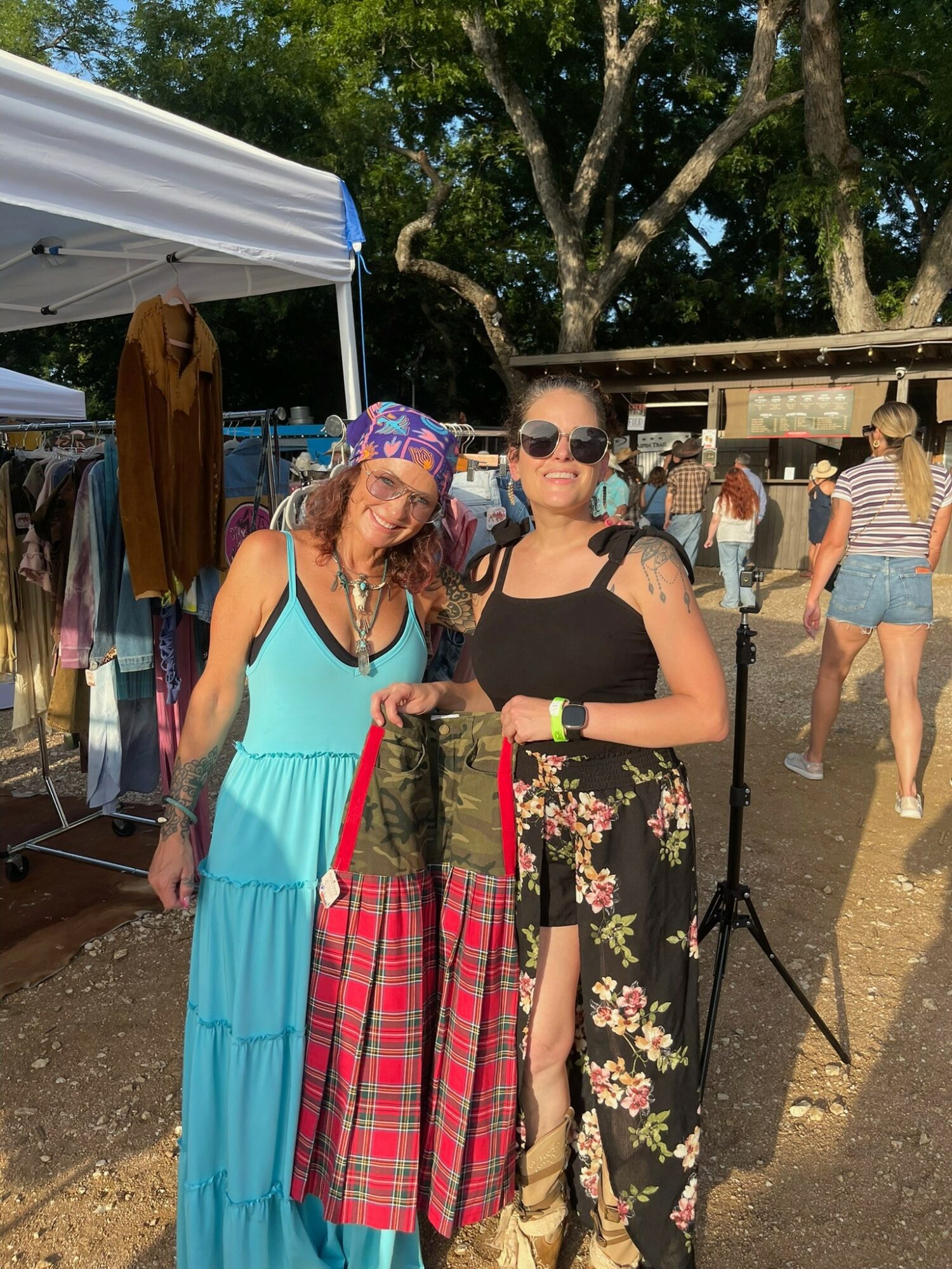 Two women standing outdoors at a market, one in a turquoise dress and the other in black with floral pants, holding a red plaid bag.