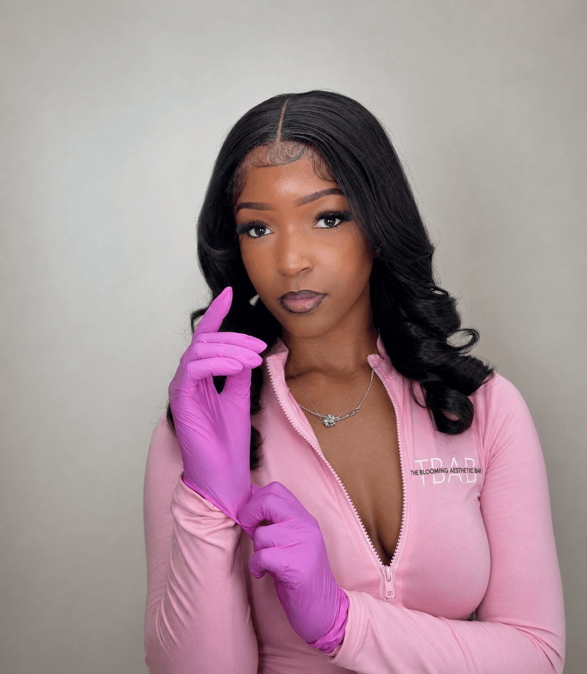 Young woman with long black hair wearing pink zip-up top and purple gloves, posing with her hand near her face.