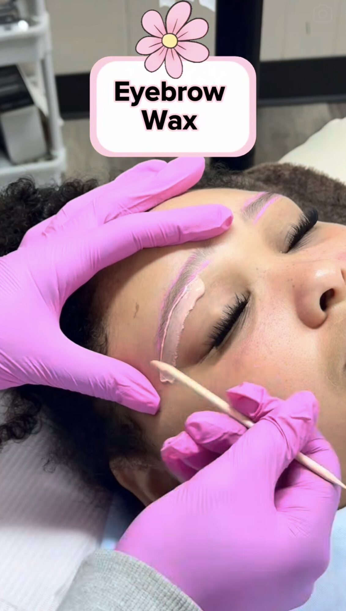 Close-up of a woman receiving eyebrow waxing, with a technician wearing pink gloves applying wax near her eyebrows.