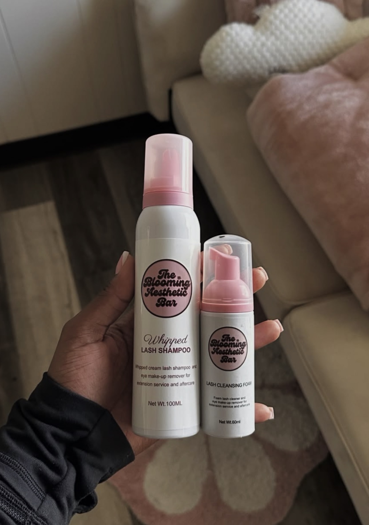 Hand holding two pink and white spray bottles with labels, in a room with a sofa and patterned rug.