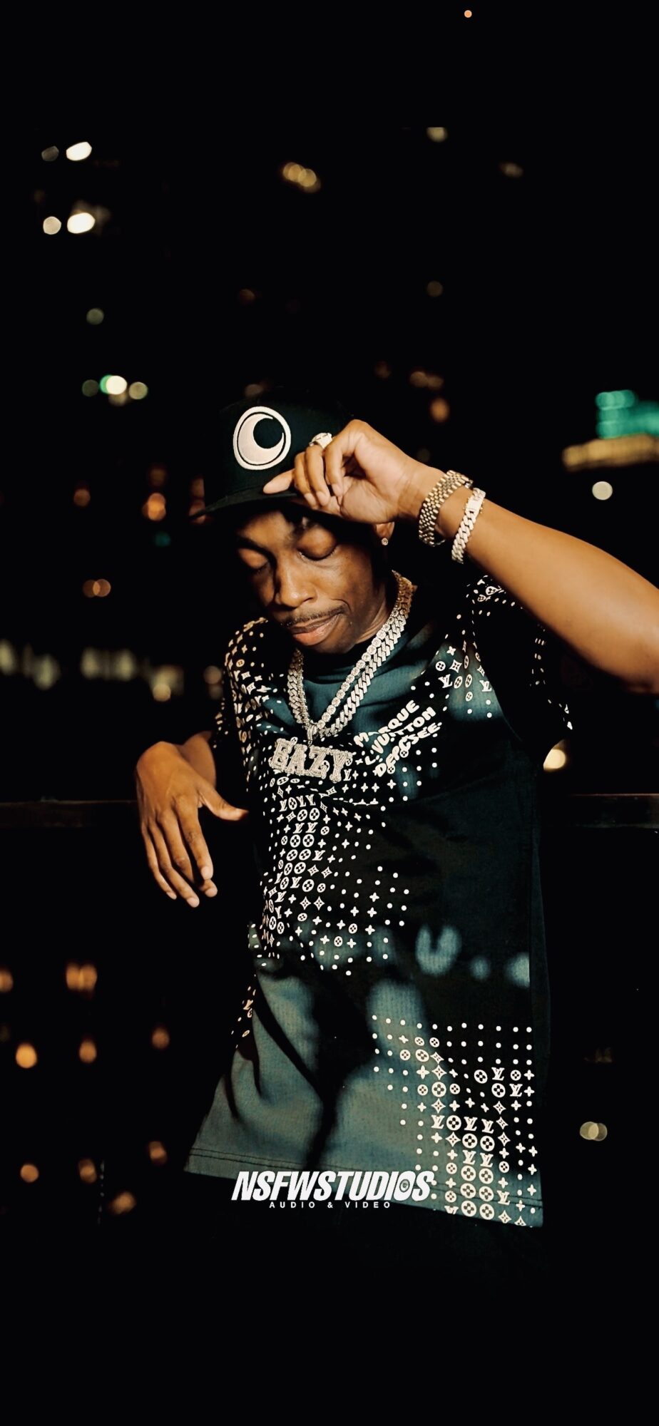 Person adjusting headphones in a city at night, wearing jewelry and a patterned shirt, with city lights in background.