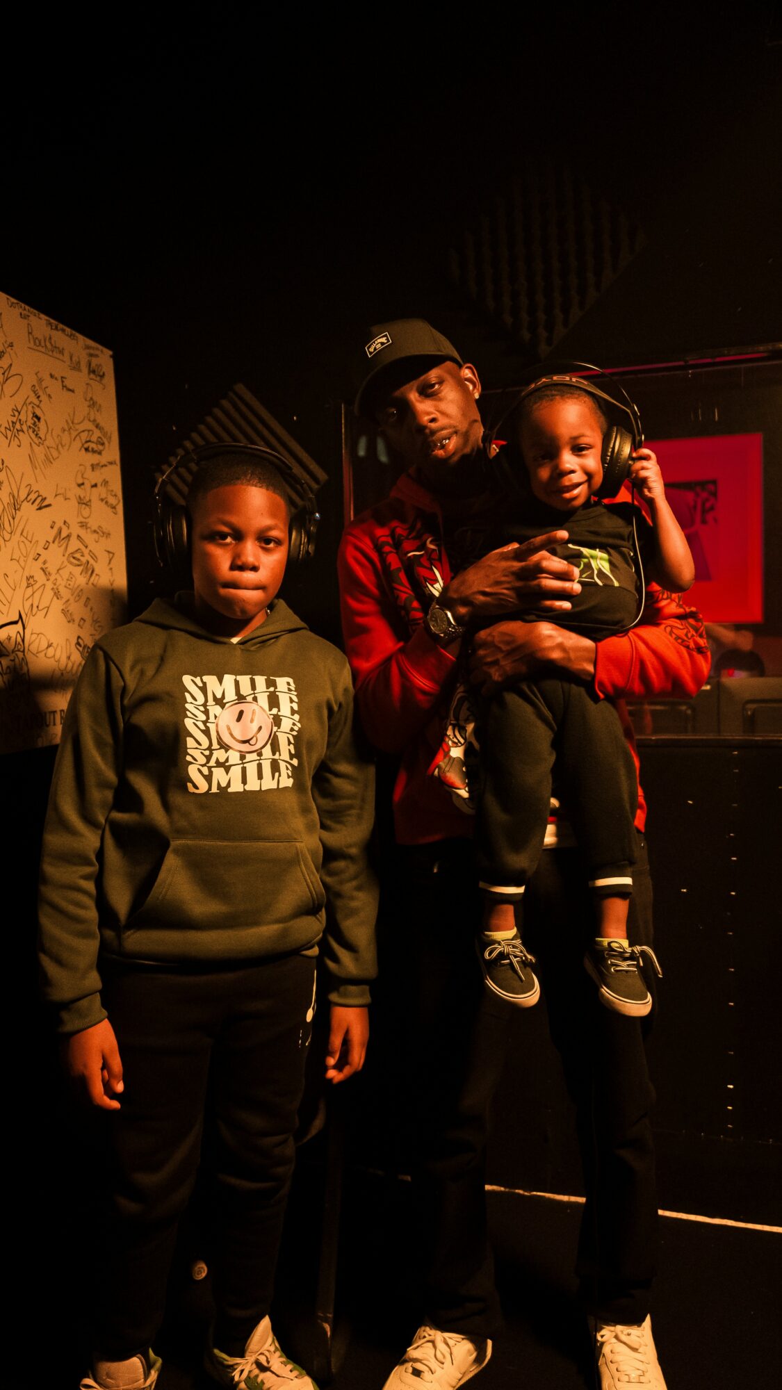 Three people in a dark room, one holding a child, all wearing casual clothing and headphones.