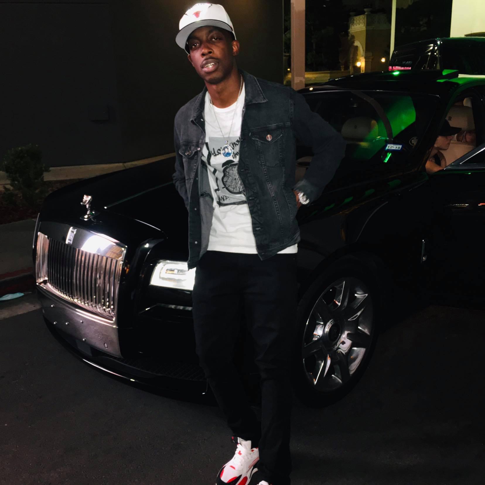 Young man standing in front of a black luxury car at night, wearing a white cap, denim jacket, and white sneakers.
