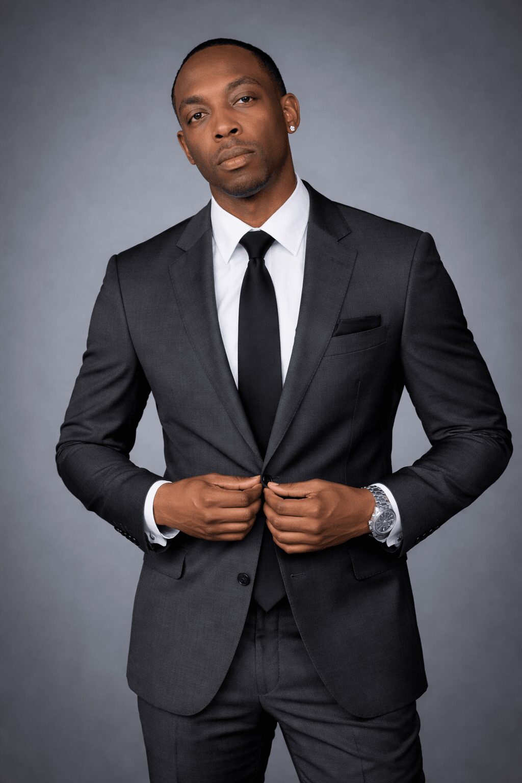 Man in a black suit, white shirt, and black tie, standing against a gray background, adjusting his suit jacket.