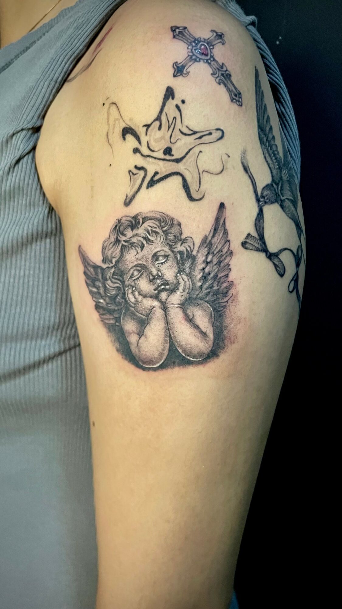Tattoo of a cherub with wings and hands on cheeks, and a cross above, on upper arm.