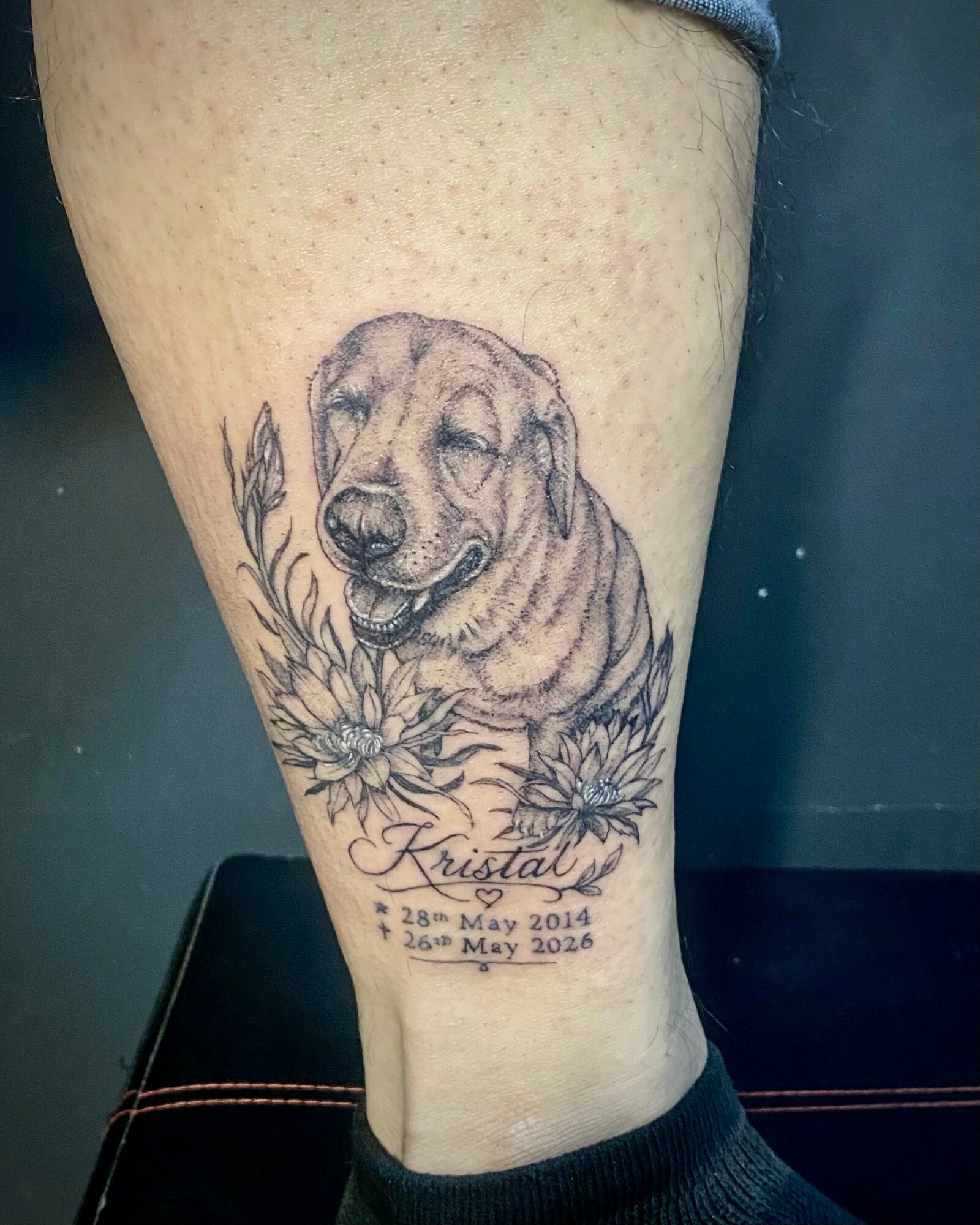 Tattoo of a smiling dog with flowers and text, including dates and a name, on a person's leg.