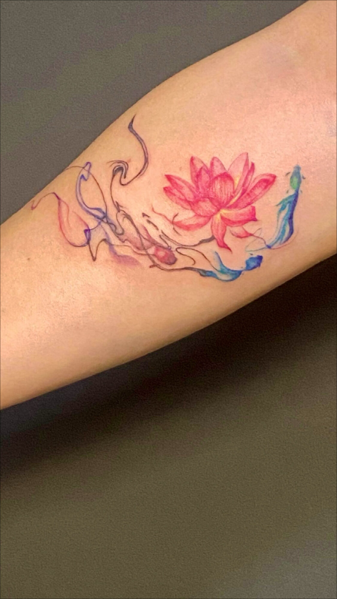Watercolor-style tattoo of a pink lotus flower with blue and purple abstract accents on skin.