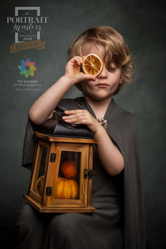 Child with curly hair holds a dried orange slice over one eye, holding a lantern with pumpkins inside, against a dark background.
