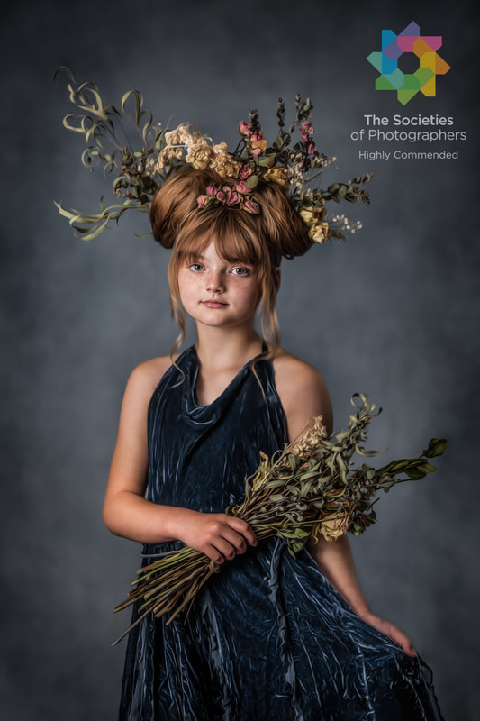 Young girl with a floral headpiece and dark dress holding a bouquet of dried flowers, against a dark background.