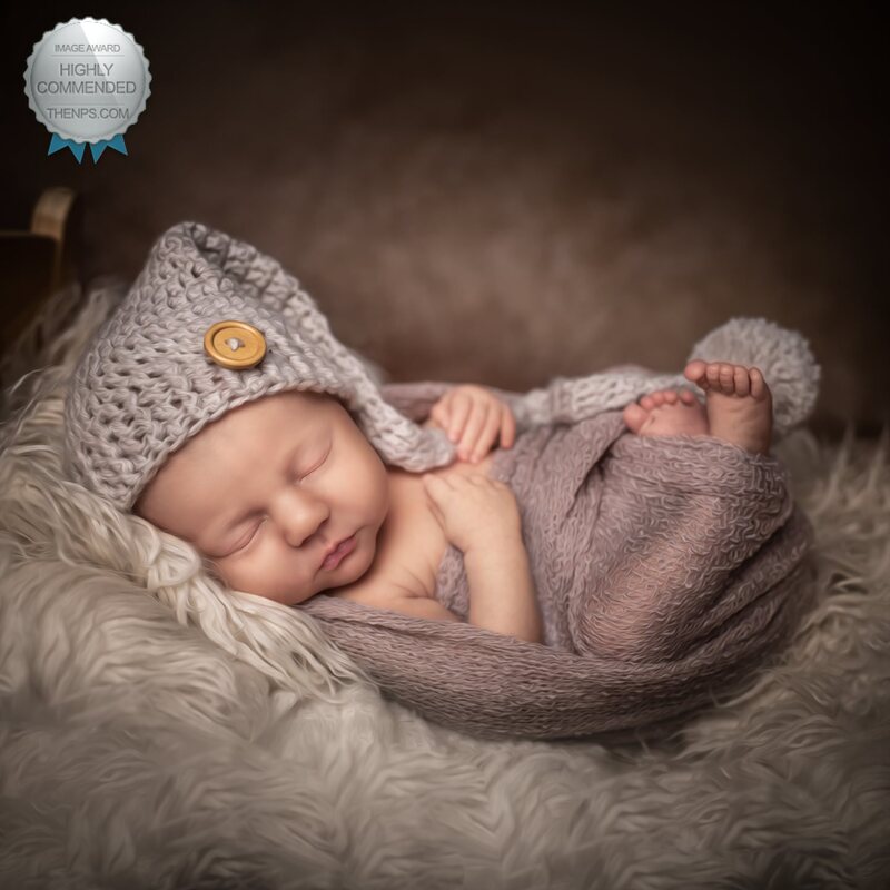 Baby sleeping on a furry surface, wearing a knitted hat with a pom-pom, wrapped in a blanket.