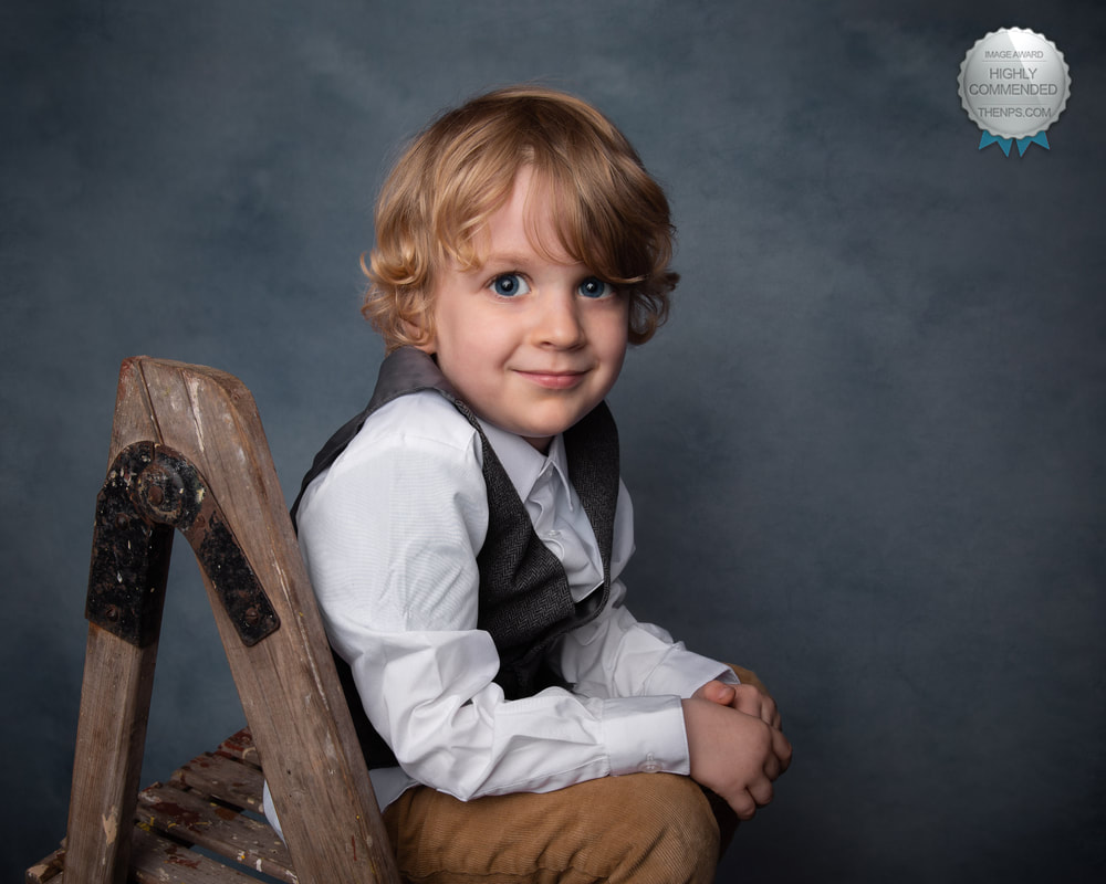 Young boy with curly blonde hair sitting on a wooden ladder, wearing a white shirt and gray vest, smiling.
