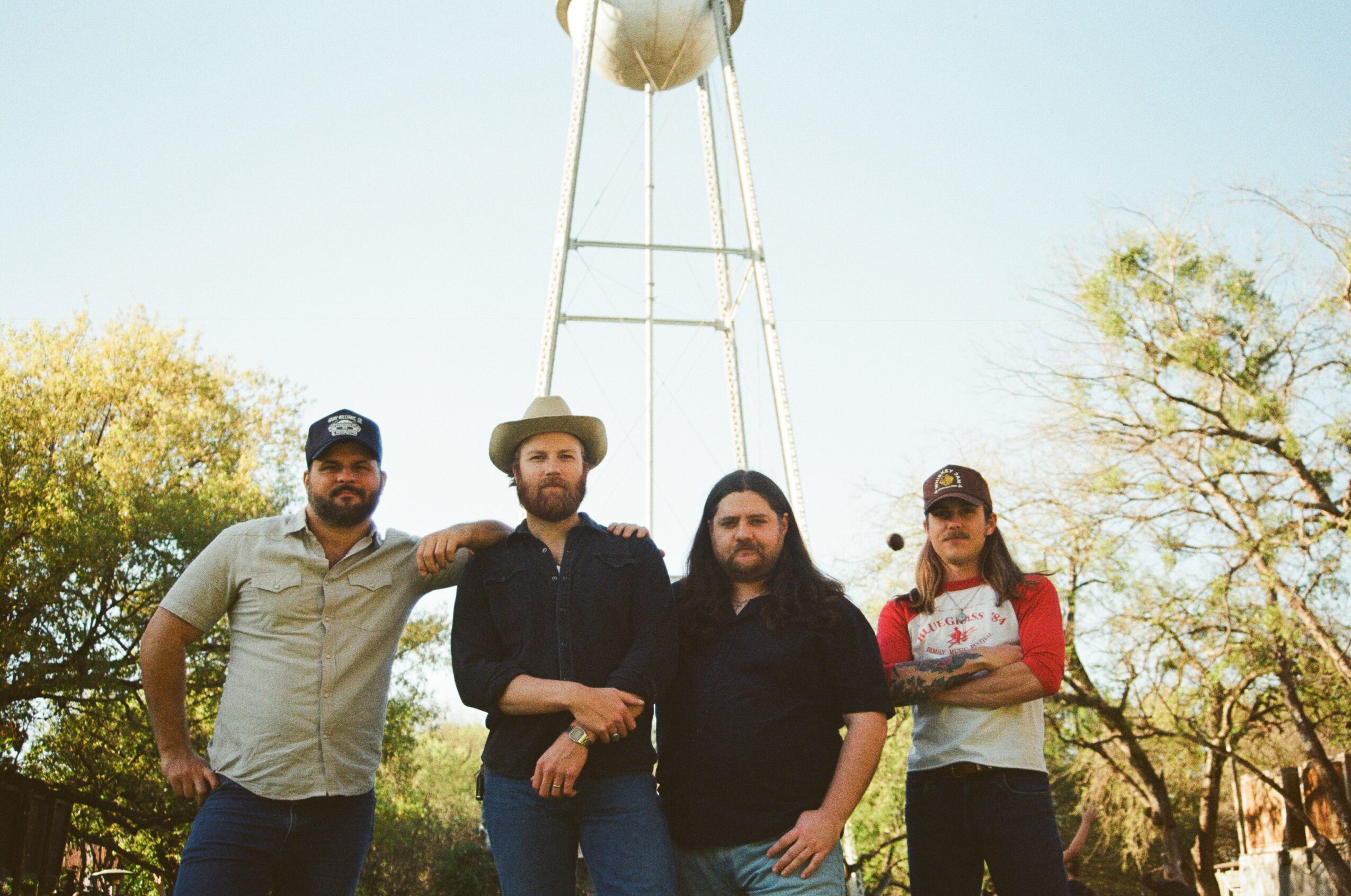Four people standing outdoors with a water tower in the background, trees, and clear sky.