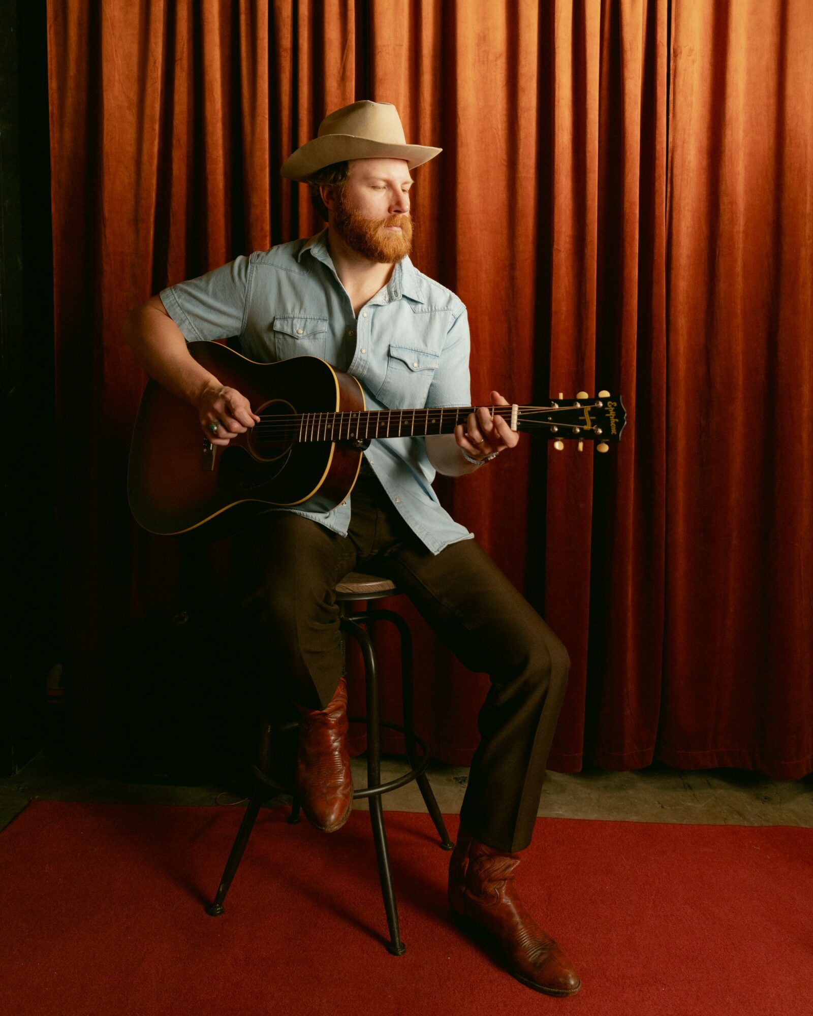 Man with a beard wearing a hat, playing guitar on a stool in front of a red curtain.