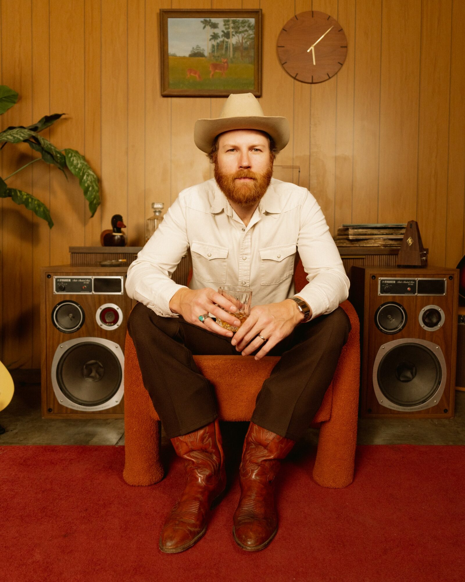 Man with a beard wearing a hat, white shirt, and brown boots sitting on a chair in a room with wood-paneled walls, speakers, and a clock.