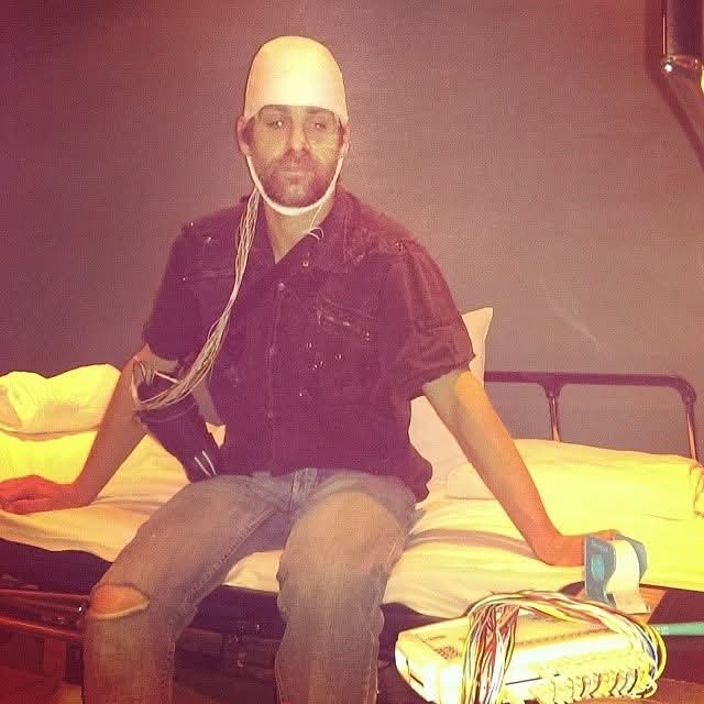 Person with a bald cap and beard sitting on a hospital bed, wearing a dark shirt and ripped jeans, with medical equipment nearby.