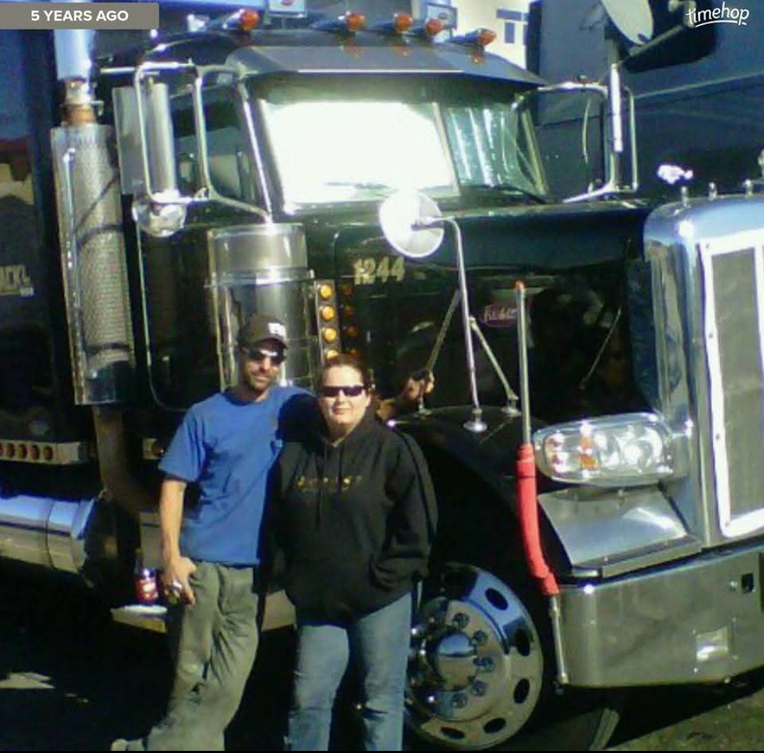 Two people stand in front of a large black semi-truck with chrome details and a red ribbon on the front, outdoors.