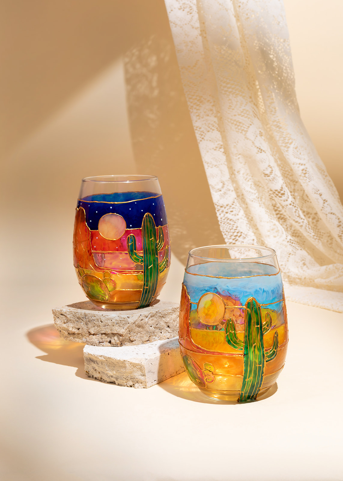 Two colorful glass cups with desert scene, cactus, and sunset, placed on stone slabs with a sheer curtain in background.