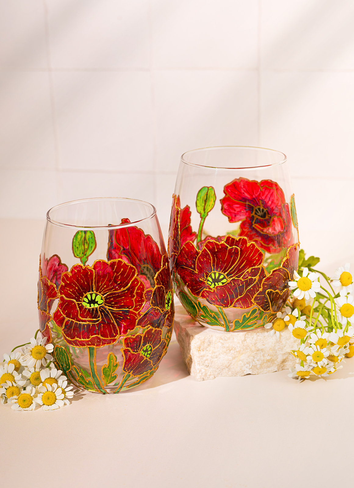 Two glasses with red and yellow floral designs, surrounded by white daisies and yellow flowers, against a white background.