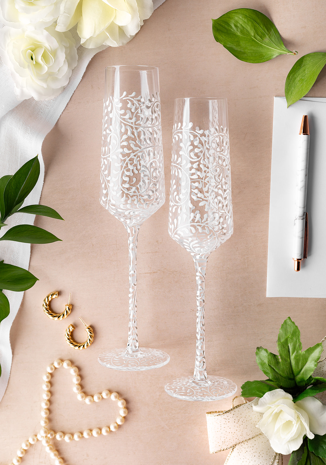 Two decorated champagne flutes on a pink surface with flowers, leaves, a pen, and a heart-shaped pearl necklace.