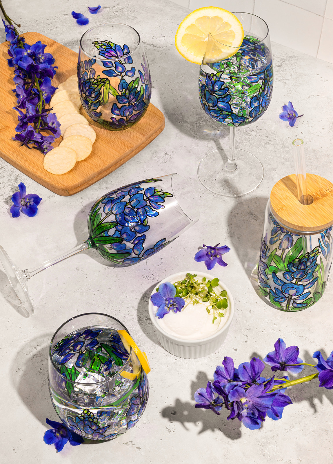 Six glasses with blue floral designs, one with lemon slice, one with wooden stick, surrounded by purple flowers on a white surface.