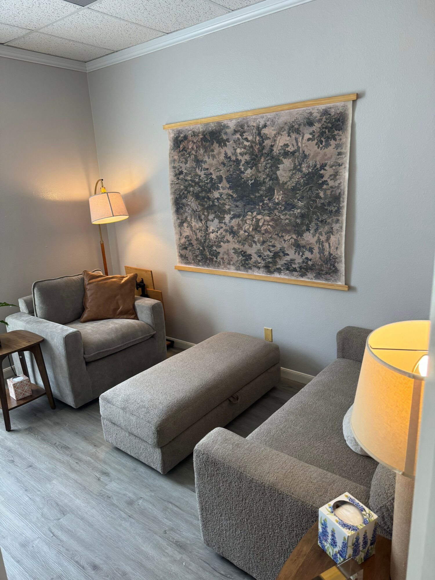Living room with a gray sofa, a wall-mounted lamp, and a textured wall hanging, viewed from the side.