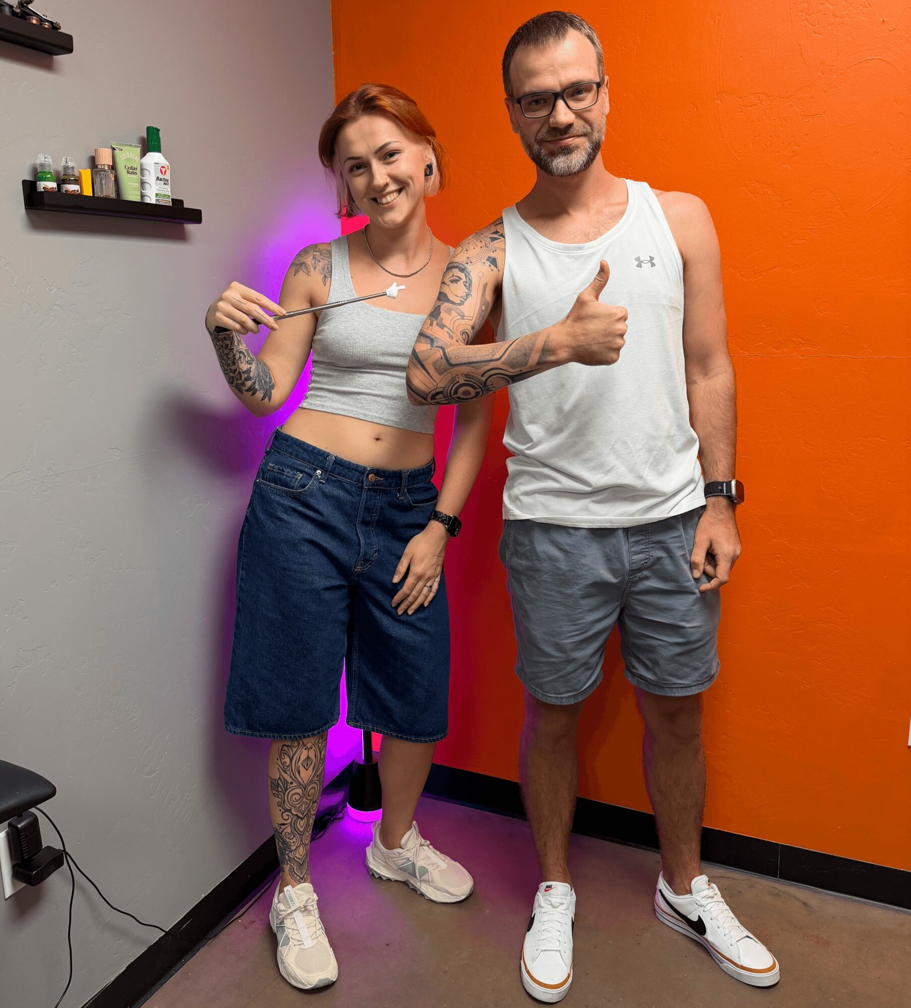 Two people, a woman and a man, stand indoors against a gray and orange wall, smiling and giving a thumbs-up. The woman has tattoos and holds a stick, wearing a crop top and shorts. The man wears a tank top and shorts. Both wear sneakers.