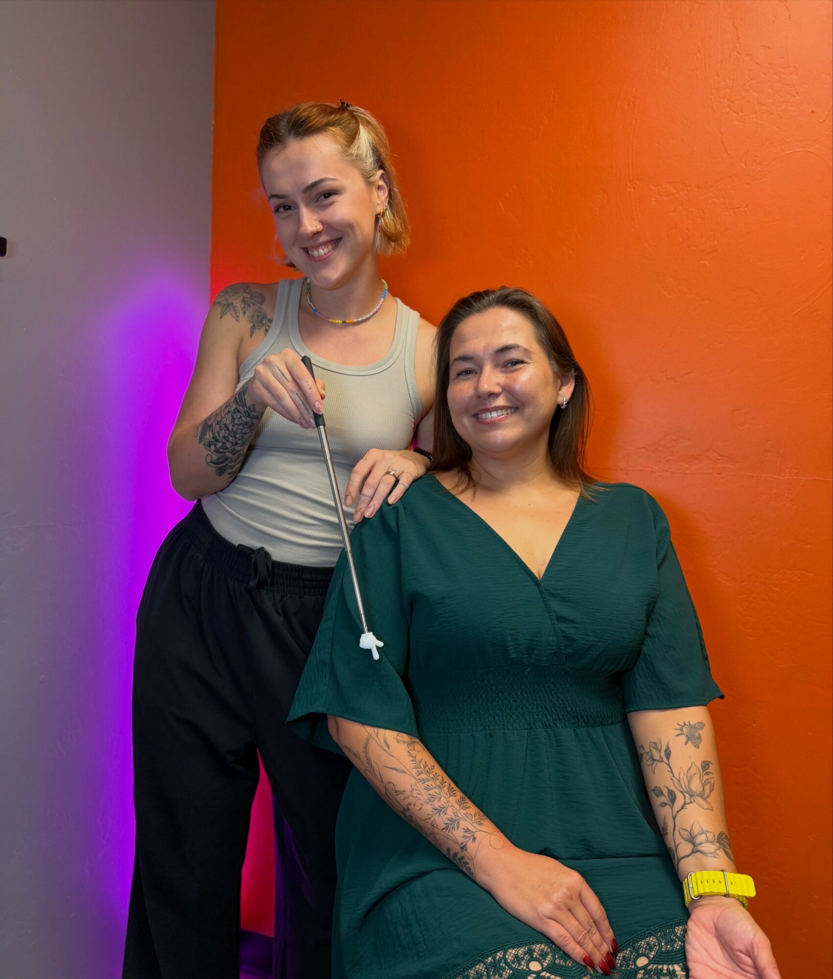 Two women smiling, one standing and one sitting, against an orange wall with purple lighting. The standing woman has blonde hair, the seated woman has dark hair and tattoos on her arms.