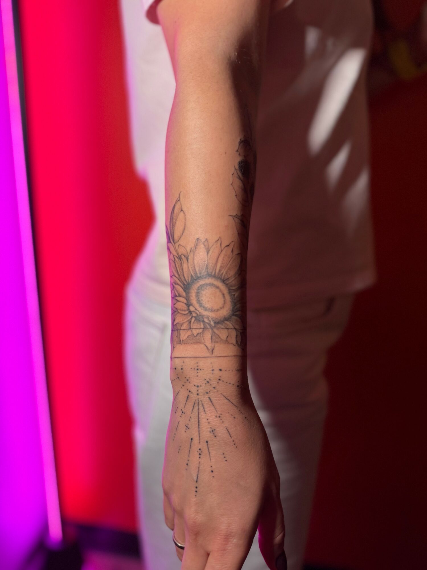 Person's arm with a tattoo of a sun and rays, standing against a colorful background.