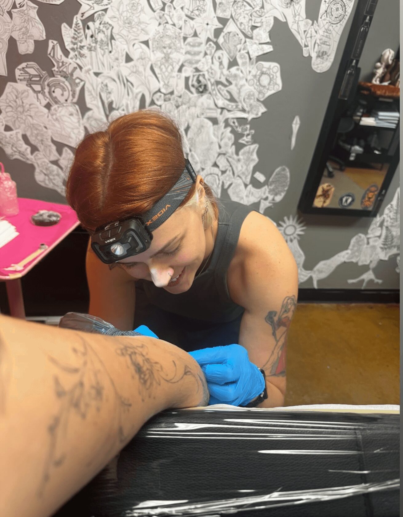 Person with red hair, wearing a headlamp and blue gloves, tattooed arm, smiling while tattooing another person's arm.