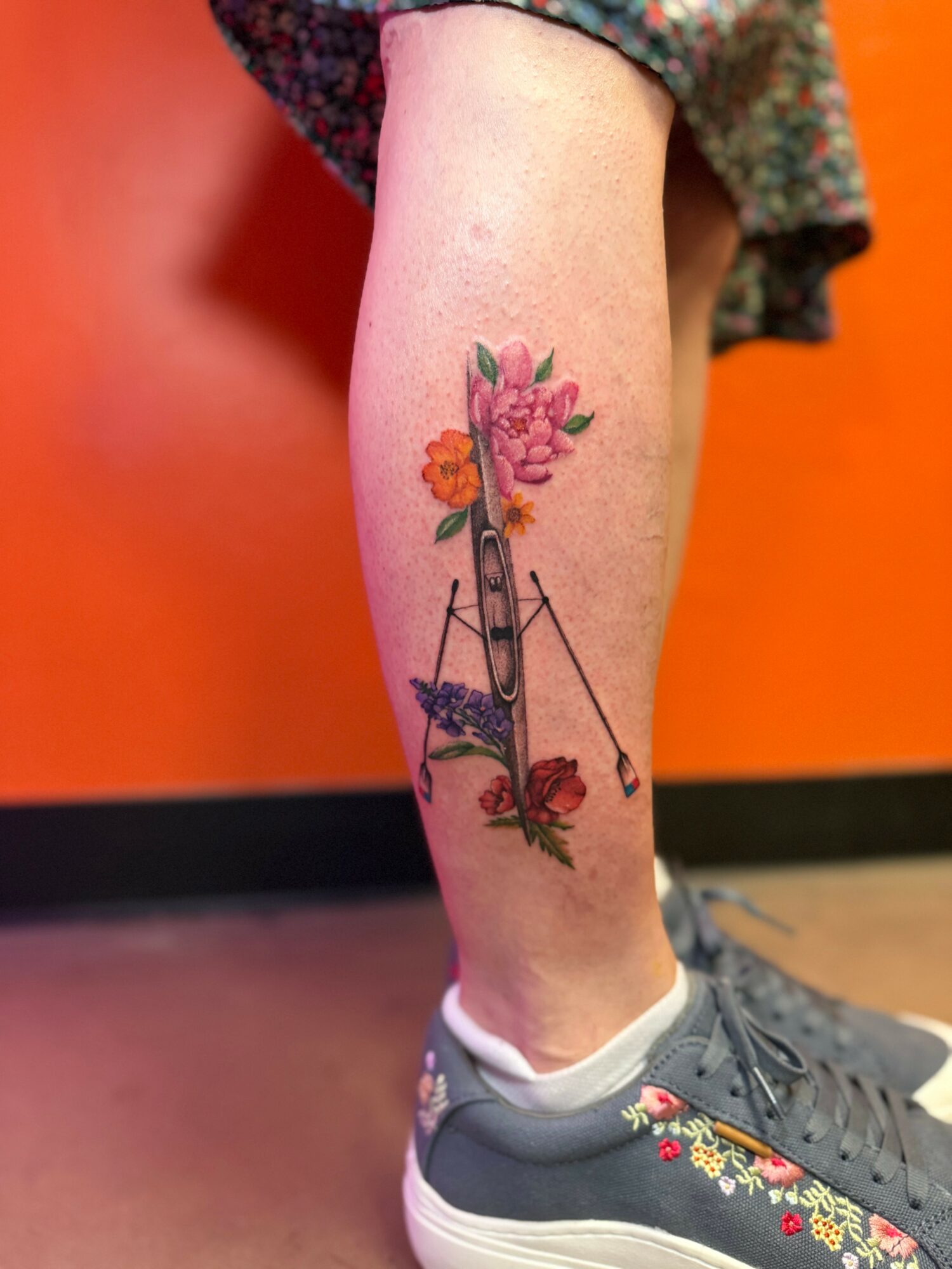 Tattoo of colorful flowers and a large safety pin on a person's lower leg, with a background of orange wall.