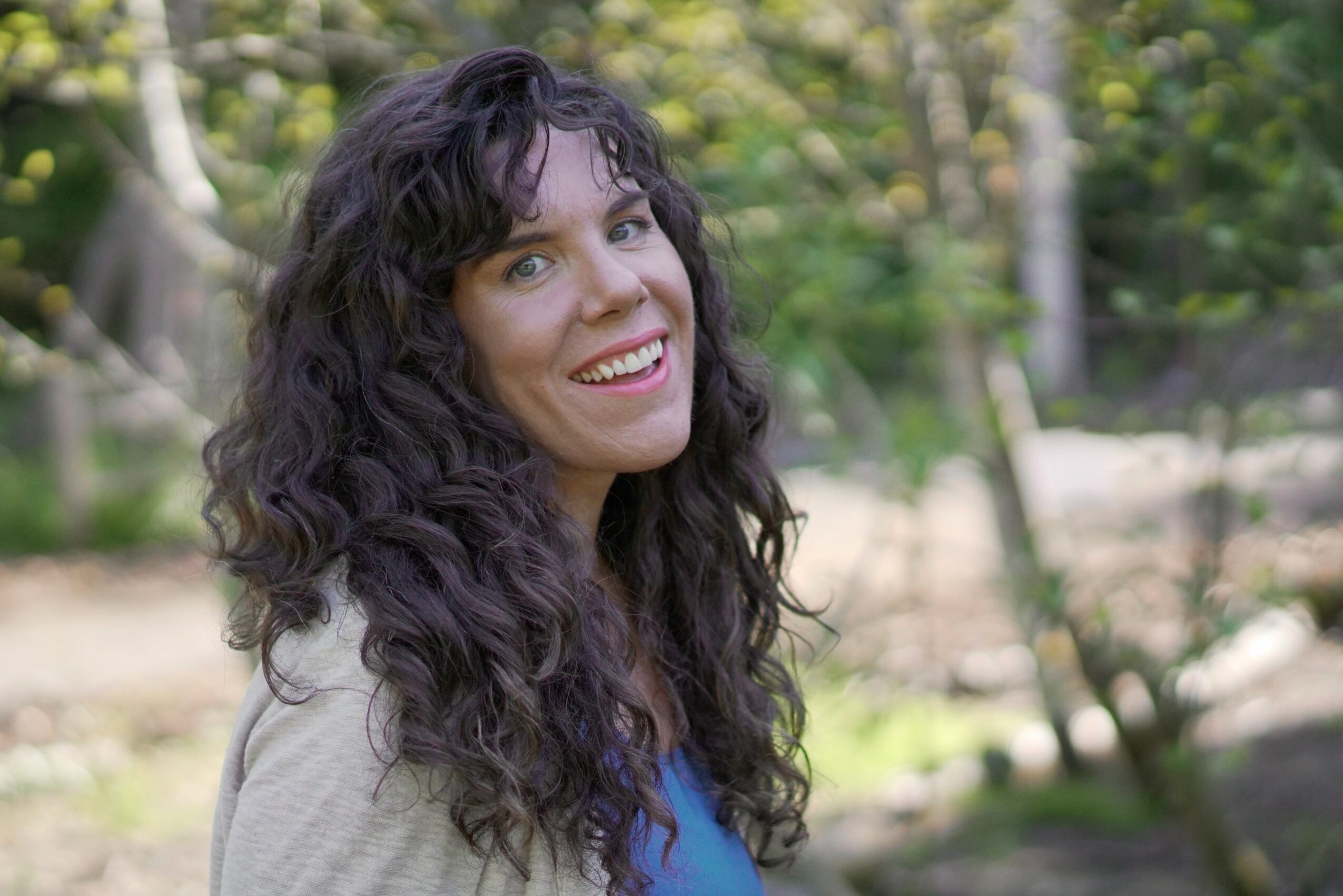 Woman with curly dark hair smiling outdoors in a park with trees and greenery