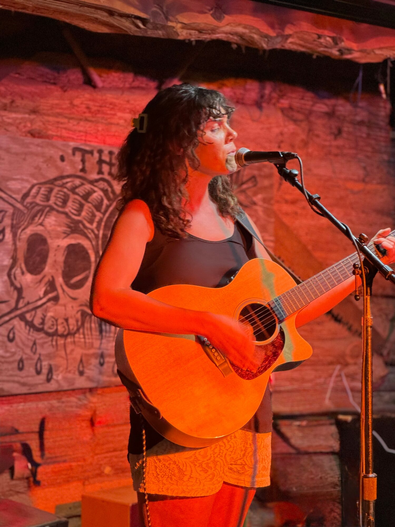 Woman with curly hair playing an acoustic guitar and singing into a microphone on stage with a skull mural background.