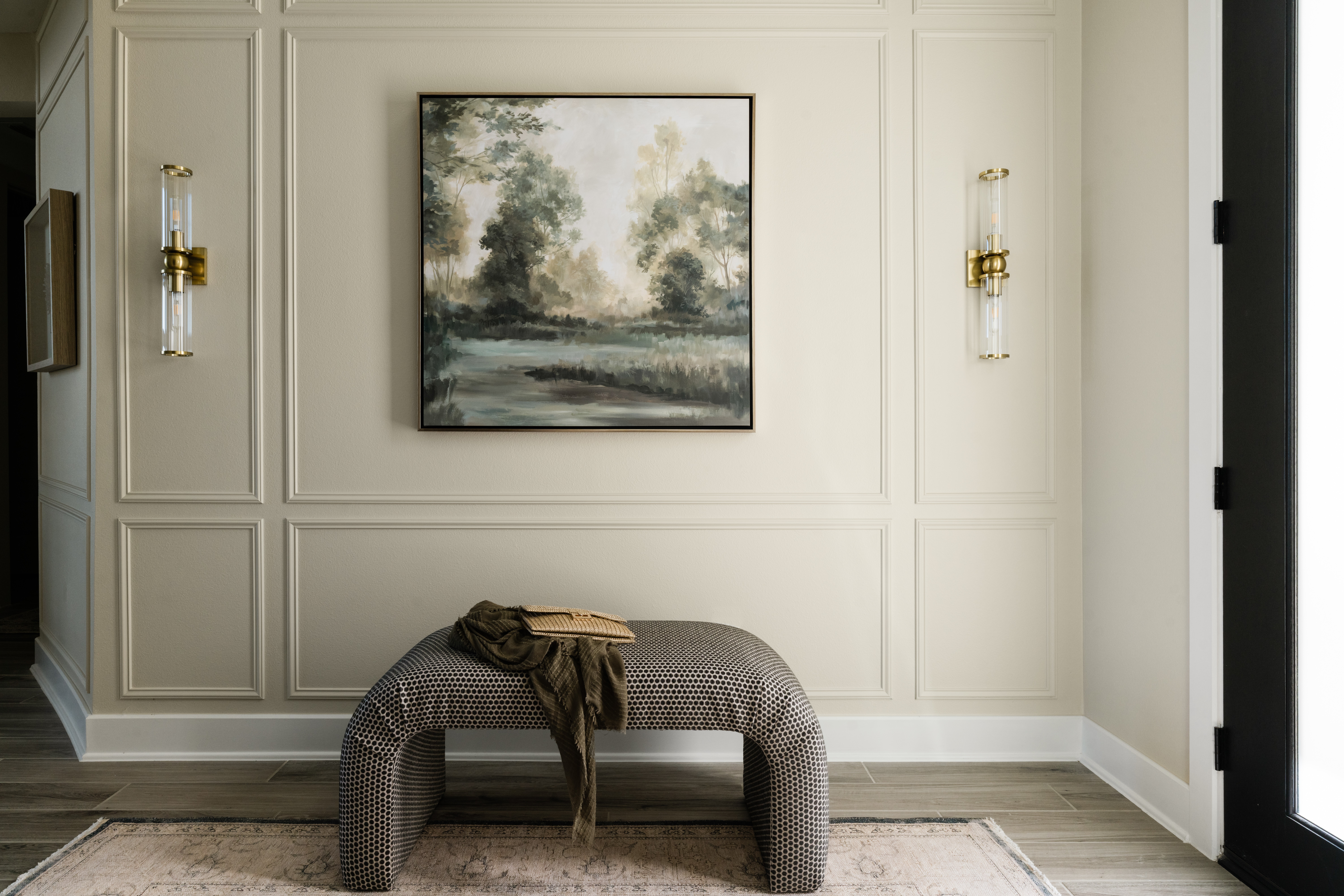 Living room with a framed landscape painting, wall sconces, a gray upholstered bench, and a window with natural light.