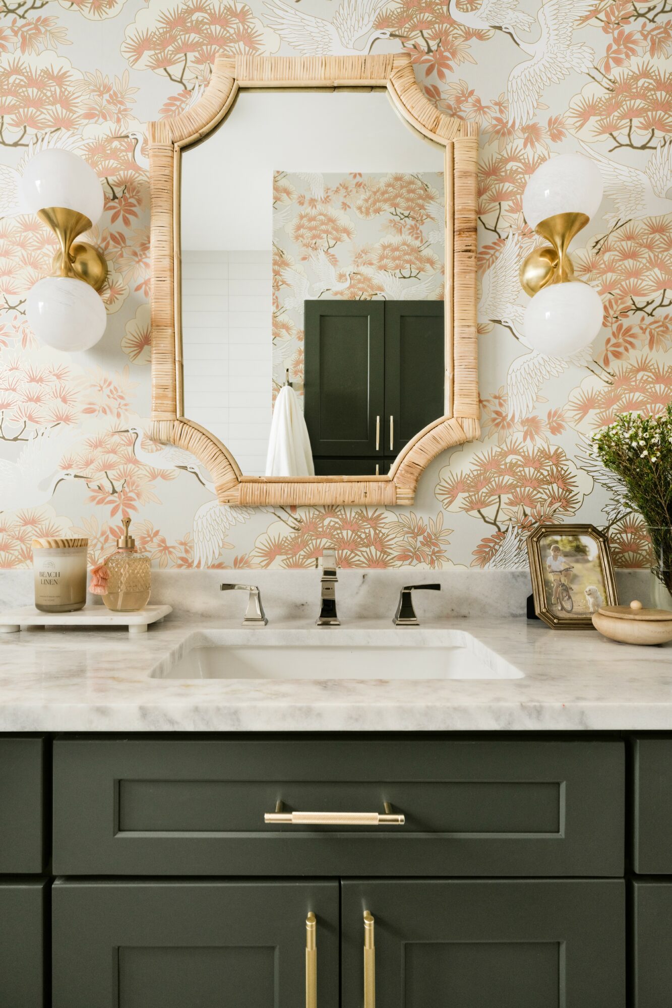 Bathroom mirror above sink, floral wallpaper, two wall sconces, countertop with decor, dark green cabinet, gold handles.