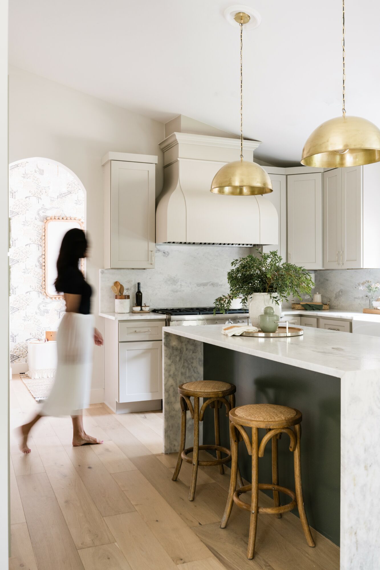 Woman walking in a modern kitchen with white cabinets, marble countertops, and gold pendant lights, with a potted plant on the island.