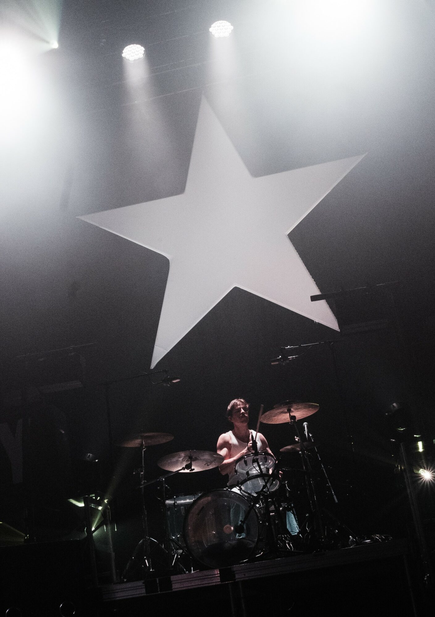 A person playing drums on stage with a large star-shaped decoration above, illuminated by stage lights.