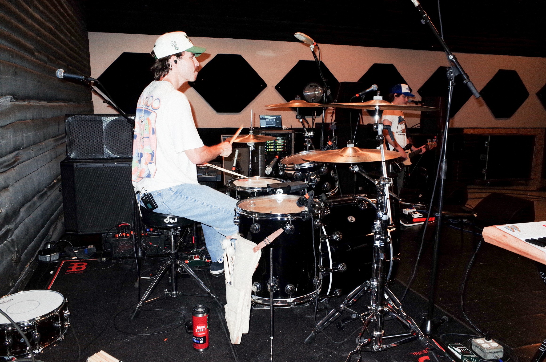 Two people playing drums in a music studio with microphones and equipment, one wearing a cap and glasses, the other with dark hair.