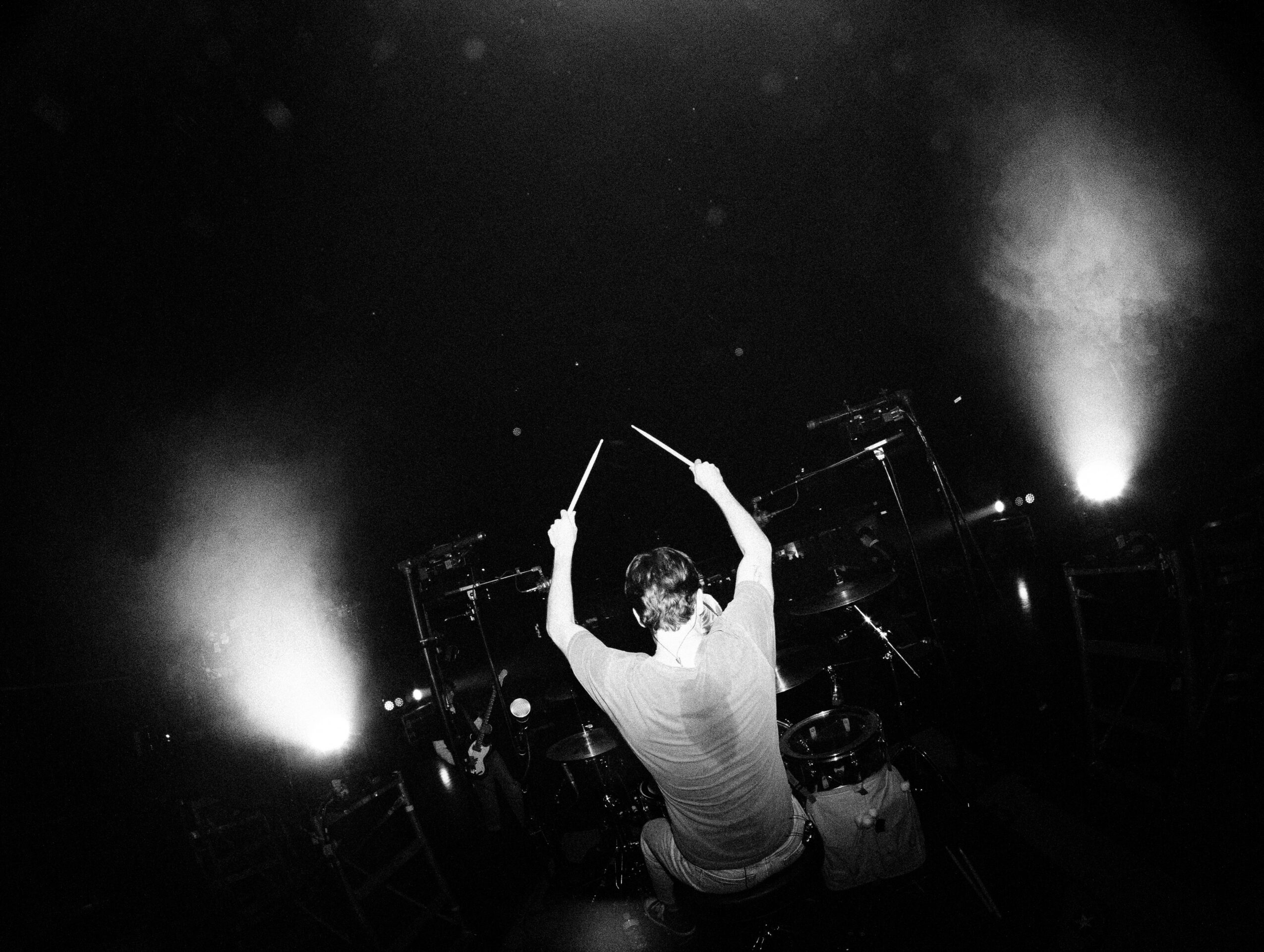Person conducting on stage with lights and equipment, back to camera, arms raised holding drumsticks, in a dark setting.